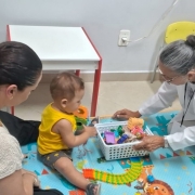 Uma fotografia colorida em plano m&eacute;dio e perspectiva em &acirc;ngulo levemente elevado regista uma sess&atilde;o de atendimento especializado infantil do programa AcompanhaRAPS em Iju&iacute;.

**Os Participantes e a A&ccedil;&atilde;o (em primeiro e meio plano):**

* No centro da composi&ccedil;&atilde;o, sobre um tapete infantil ilustrado e colorido em tons azuis com desenhos de animais e trilhos, tr&ecirc;s pessoas est&atilde;o sentadas ao n&iacute;vel do ch&atilde;o:


* &Agrave; esquerda, em primeiro plano, uma mulher de costas e perfil de cabelos escuros apanhados num coque veste camisola bege de malha texturizada e acompanha o filho.


* No centro, um beb&eacute; de camisa amarela sem mangas e cal&ccedil;&otilde;es escuros brinca no ch&atilde;o com blocos pedag&oacute;gicos de encaixe e brinquedos coloridos.


* &Agrave; direita, uma profissional de sa&uacute;de de cabelos grisalhos apanhados, vestindo bata/jaleco branco sobre roupa clara e crach&aacute; ao pesco&ccedil;o, est&aacute; ajoelhada e aproxima com cuidado um cesto pl&aacute;stico branco repleto de brinquedos pedag&oacute;gicos para a crian&ccedil;a.

**O Ambiente e o Mobili&aacute;rio (ao fundo):**

* Ao fundo, v&ecirc;-se uma pequena mesa infantil quadrada com tampo branco e pernas vermelhas.

* Ao lado da mesa, observam-se duas cadeiras infantis em tons de verde-menta e amarelo.

* As paredes da sala de atendimento s&atilde;o claras e lisas, com tomada el&eacute;trica vis&iacute;vel e ilumina&ccedil;&atilde;o interior direta e acolhedora.