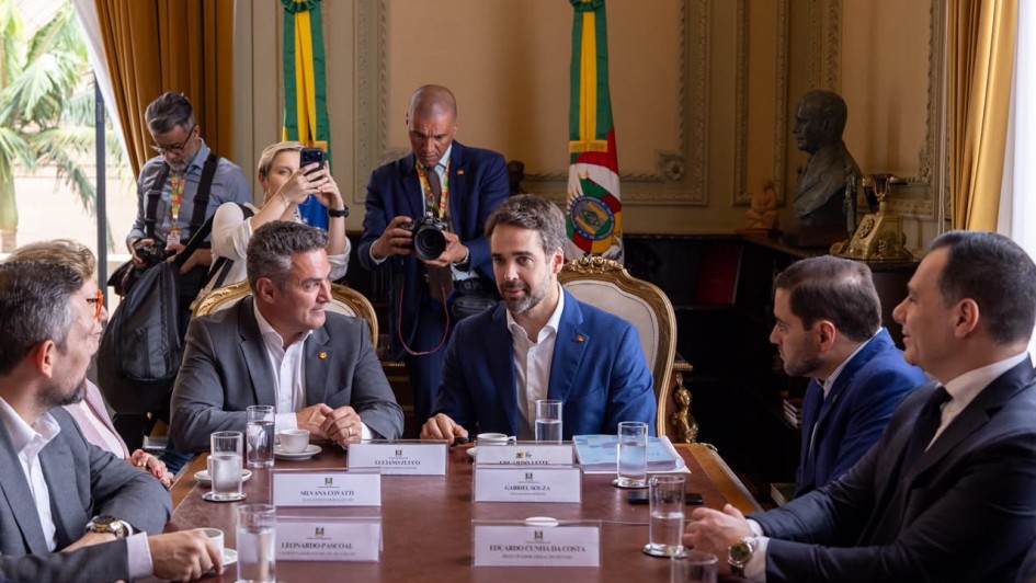 Uma fotografia colorida em plano m&eacute;dio-geral e perspectiva ao n&iacute;vel dos olhos regista a primeira reuni&atilde;o oficial de transi&ccedil;&atilde;o de governo entre o governador Eduardo Leite e o governador eleito Luciano Zucco, acompanhados por membros das respetivas equipas, no Pal&aacute;cio Piratini.

**As Autoridades e a Mesa de Reuni&atilde;o (ao centro e em primeiro plano):**

* Ao centro da mesa de madeira nobre, o governador Eduardo Leite fala de forma atenta, vestindo fato azul-vibrante, camisa branca sem gravata e pin institucional na lapela.
* &Agrave; sua esquerda (&agrave; direita do observador), Luciano Zucco (governador eleito) escuta com aten&ccedil;&atilde;o, trajando fato cinzento-escuro e camisa branca sem gravata.
* Ladeando a mesa retangular, alinham-se outros integrantes das equipas de transi&ccedil;&atilde;o com identificadores nominais em acr&iacute;lico &agrave; frente de cada lugar (como Gabriel Souza, Silvana Covatti, Leonardo Pascoal e Eduardo Cunha da Costa):
* Em primeiro plano, &agrave; direita, sobressaem-se autoridades em fatos escuros e gravatas formais.
* &Agrave; esquerda, veem-se membros da equipa sentados, com copos de &aacute;gua e x&iacute;caras de caf&eacute; dispostos sobre a bancada.



**A Imprensa e o Ambiente de Fundo (ao fundo):**

* Diretamente atr&aacute;s do governador e do governador eleito, em p&eacute;, tr&ecirc;s profissionais de imprensa e fot&oacute;grafos registam o encontro com c&acirc;mara fotogr&aacute;fica profissional e smartphone.
* Ao fundo, adornando o sal&atilde;o nobre, destacam-se duas grandes bandeiras oficiais em mastros (a bandeira do Estado do Rio Grande do Sul e a bandeira do Brasil com la&ccedil;o de ins&iacute;gnia).
* A sala exibe arquitetura cl&aacute;ssica com paredes em tom creme, bustos esculpidos e cortinados em tom dourado, sob ilumina&ccedil;&atilde;o interior quente e uniforme.