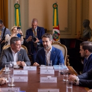 Uma fotografia colorida em plano m&eacute;dio-geral e perspectiva ao n&iacute;vel dos olhos regista a primeira reuni&atilde;o oficial de transi&ccedil;&atilde;o de governo entre o governador Eduardo Leite e o governador eleito Luciano Zucco, acompanhados por membros das respetivas equipas, no Pal&aacute;cio Piratini.

**As Autoridades e a Mesa de Reuni&atilde;o (ao centro e em primeiro plano):**

* Ao centro da mesa de madeira nobre, o governador Eduardo Leite fala de forma atenta, vestindo fato azul-vibrante, camisa branca sem gravata e pin institucional na lapela.
* &Agrave; sua esquerda (&agrave; direita do observador), Luciano Zucco (governador eleito) escuta com aten&ccedil;&atilde;o, trajando fato cinzento-escuro e camisa branca sem gravata.
* Ladeando a mesa retangular, alinham-se outros integrantes das equipas de transi&ccedil;&atilde;o com identificadores nominais em acr&iacute;lico &agrave; frente de cada lugar (como Gabriel Souza, Silvana Covatti, Leonardo Pascoal e Eduardo Cunha da Costa):
* Em primeiro plano, &agrave; direita, sobressaem-se autoridades em fatos escuros e gravatas formais.
* &Agrave; esquerda, veem-se membros da equipa sentados, com copos de &aacute;gua e x&iacute;caras de caf&eacute; dispostos sobre a bancada.



**A Imprensa e o Ambiente de Fundo (ao fundo):**

* Diretamente atr&aacute;s do governador e do governador eleito, em p&eacute;, tr&ecirc;s profissionais de imprensa e fot&oacute;grafos registam o encontro com c&acirc;mara fotogr&aacute;fica profissional e smartphone.
* Ao fundo, adornando o sal&atilde;o nobre, destacam-se duas grandes bandeiras oficiais em mastros (a bandeira do Estado do Rio Grande do Sul e a bandeira do Brasil com la&ccedil;o de ins&iacute;gnia).
* A sala exibe arquitetura cl&aacute;ssica com paredes em tom creme, bustos esculpidos e cortinados em tom dourado, sob ilumina&ccedil;&atilde;o interior quente e uniforme.