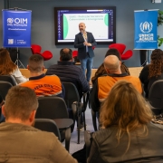 Foto mostra um palestrante, usando um blazer cinza, fala diante da plateia formada por homens e mulheres. Alguns deles usam colete laranja da Defesa Civil.