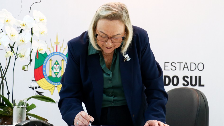 Uma fotografia colorida em plano m&eacute;dio-fechado e perspectiva ao n&iacute;vel dos olhos registra o momento em que a secret&aacute;ria estadual da Sa&uacute;de, Arita Bergmann, assina um conv&ecirc;nio oficial.

**A Secret&aacute;ria e o Ato de Assinatura (ao centro):**

* No centro da composi&ccedil;&atilde;o, a secret&aacute;ria Arita Bergmann aparece inclinada sobre uma bancada clara enquanto assina um documento com uma caneta branca na m&atilde;o direita.
* Ela possui cabelos loiros/grisalhos m&eacute;dios &agrave; altura dos ombros, usa &oacute;culos de arma&ccedil;&atilde;o escura e veste um blazer azul-marinho sobre uma camisa social verde-escura.
* Na lapela do blazer, destaca-se um broche prateado com pedras em formato de flor.
* Sobre a bancada, &agrave; esquerda, observa-se um vaso decorativo com orqu&iacute;deas brancas e um copo de &aacute;gua.

**O Painel de Fundo (ao fundo):**

* Ao fundo, a parede branca exibe elementos da identidade visual do Governo do Estado do Rio Grande do Sul.
* &Agrave; esquerda, destaca-se o bras&atilde;o de armas oficial do Estado do Rio Grande do Sul impresso em cores vivas (verde, vermelho e amarelo).
* &Agrave; direita, l&ecirc;-se parcialmente em letras garrafais pretas mai&uacute;sculas o texto: **"ESTADO DO Rio Grande do SUL"**.
* A ilumina&ccedil;&atilde;o no ambiente &eacute; direta, uniforme e bem distribu&iacute;da.

---