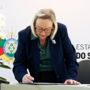 Uma fotografia colorida em plano m&eacute;dio-fechado e perspectiva ao n&iacute;vel dos olhos registra o momento em que a secret&aacute;ria estadual da Sa&uacute;de, Arita Bergmann, assina um conv&ecirc;nio oficial.

**A Secret&aacute;ria e o Ato de Assinatura (ao centro):**

* No centro da composi&ccedil;&atilde;o, a secret&aacute;ria Arita Bergmann aparece inclinada sobre uma bancada clara enquanto assina um documento com uma caneta branca na m&atilde;o direita.
* Ela possui cabelos loiros/grisalhos m&eacute;dios &agrave; altura dos ombros, usa &oacute;culos de arma&ccedil;&atilde;o escura e veste um blazer azul-marinho sobre uma camisa social verde-escura.
* Na lapela do blazer, destaca-se um broche prateado com pedras em formato de flor.
* Sobre a bancada, &agrave; esquerda, observa-se um vaso decorativo com orqu&iacute;deas brancas e um copo de &aacute;gua.

**O Painel de Fundo (ao fundo):**

* Ao fundo, a parede branca exibe elementos da identidade visual do Governo do Estado do Rio Grande do Sul.
* &Agrave; esquerda, destaca-se o bras&atilde;o de armas oficial do Estado do Rio Grande do Sul impresso em cores vivas (verde, vermelho e amarelo).
* &Agrave; direita, l&ecirc;-se parcialmente em letras garrafais pretas mai&uacute;sculas o texto: **"ESTADO DO Rio Grande do SUL"**.
* A ilumina&ccedil;&atilde;o no ambiente &eacute; direta, uniforme e bem distribu&iacute;da.

---