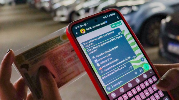Foto mostra uma pessoa segurando um celular com a tela aberta no Whatsapp na conversa com a assistente Guria. Na outra m&atilde;o, a pessoa segura uma carteira de motorista CNH. Ao fundo, desfocados, aparecem ve&iacute;culos num grande estacionamento. 