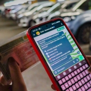 Foto mostra uma pessoa segurando um celular com a tela aberta no Whatsapp na conversa com a assistente Guria. Na outra m&atilde;o, a pessoa segura uma carteira de motorista CNH. Ao fundo, desfocados, aparecem ve&iacute;culos num grande estacionamento. 