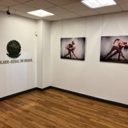 Uma fotografia colorida em plano geral e perspectiva em &acirc;ngulo levemente elevado capta a &aacute;rea de exposi&ccedil;&otilde;es no Consulado-Geral do Brasil em Nova Iorque, sem a presen&ccedil;a de pessoas.

**A Identifica&ccedil;&atilde;o Institucional (&agrave; esquerda):**

* Na parede branca do lado esquerdo, destaca-se em relevo met&aacute;lico escuro o Bras&atilde;o da Rep&uacute;blica Federativa do Brasil.


* Logo abaixo do bras&atilde;o, l&ecirc;-se a inscri&ccedil;&atilde;o em letras garrafais mai&uacute;sculas met&aacute;licas: **"CONSULADO-GERAL DO BRASIL"**.



**A Galeria e as Obras Expostas (ao fundo):**

* Na parede branca do fundo, encontram-se expostas duas fotografias art&iacute;sticas retangulares de grande formato, suspensas por finos cabos met&aacute;licos presos ao teto:


* A fotografia da esquerda retrata um casal de bailarinos numa pose de dan&ccedil;a expressiva sob jorros de &aacute;gua/chuva iluminados.


* A fotografia da direita exibe o mesmo casal numa pose din&acirc;mica de dan&ccedil;a contempor&acirc;nea, com movimentos dram&aacute;ticos e salpicos de &aacute;gua ao fundo.





**O Espa&ccedil;o e a Ilumina&ccedil;&atilde;o:**

* O espa&ccedil;o possui piso em reguado de madeira em tom castanho-m&eacute;dio com acabamento acetinado e rodap&eacute;s escuros ao longo das paredes brancas e lisas.


* O teto modular em placas brancas conta com pain&eacute;is de ilumina&ccedil;&atilde;o encastrados que emitem uma luz suave e uniforme sobre a &aacute;rea de exposi&ccedil;&atilde;o.