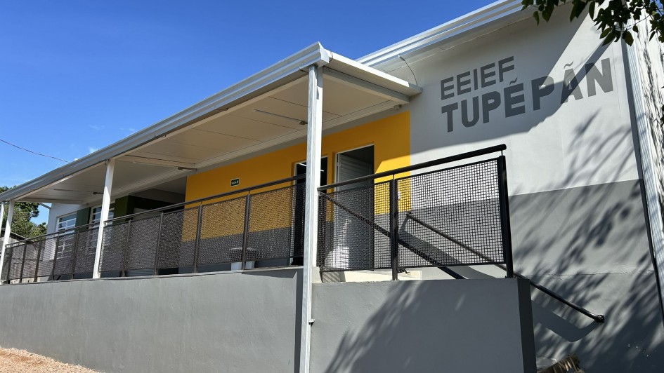 Uma fotografia colorida em plano m&eacute;dio-geral e perspectiva em contra-plong&eacute;e (de baixo para cima) capta a fachada rec&eacute;m reformada da Escola Estadual Ind&iacute;gena de Ensino Fundamental Tupẽ P&atilde;n.

**A Fachada da Escola e a Acessibilidade (ao centro e em primeiro plano):**

* No centro e &agrave; direita da composi&ccedil;&atilde;o, a parede do edif&iacute;cio exibe pintura em dois tons: cinzento-claro na metade superior e cinzento-escuro na metade inferior.


* Na parte superior direita da fachada, l&ecirc;-se em letras mai&uacute;sculas garrafais de cor cinzento-escuro a identifica&ccedil;&atilde;o da escola: **"EEIEF TUPẼ P&Atilde;N"**.


* Em primeiro plano, estende-se uma rampa e plataforma elevada com mureta cinzenta refor&ccedil;ada por um guarda-corpo met&aacute;lico com grelha de prote&ccedil;&atilde;o preta. &Agrave; direita, v&ecirc;-se uma pequena escada de acesso acompanhada por corrim&atilde;o tubular preto.



**A Varanda e a Estrutura (&agrave; esquerda e ao fundo):**

* &Agrave; esquerda, uma varanda coberta com teto inclinado e calhas brancas apoia-se em colunas met&aacute;licas brancas.


* A parede ao fundo da varanda apresenta um painel em amarelo vibrante com portas e janelas de acesso &agrave;s salas de aula.


* O solo em primeiro plano &eacute; constitu&iacute;do por terra e gravilha.


* No canto superior direito, sobressai a folhagem de uma &aacute;rvore sob um c&eacute;u azul-intenso e limpo, banhado por luz solar direta.
