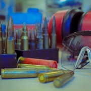 Foto mostra cartuchos de armas de fogo em estojos e espalhados sobre a bancada. Ao lado est&aacute; um &oacute;culos de prote&ccedil;&atilde;o.