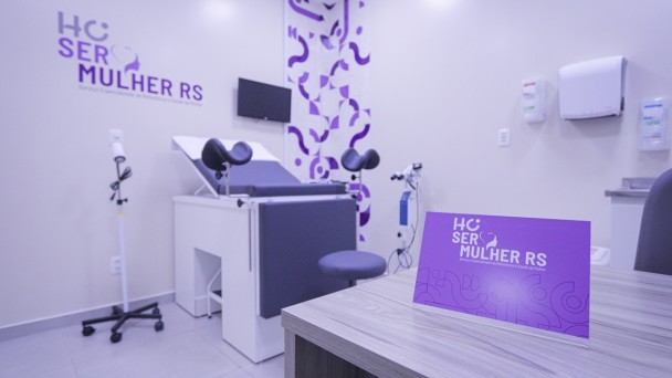 Uma fotografia colorida em plano m&eacute;dio e perspectiva em &acirc;ngulo levemente baixo capta o interior de uma sala de exames ginecol&oacute;gicos especializada e moderna do espa&ccedil;o SER Mulher RS, sem a presen&ccedil;a de pessoas.

**A Identifica&ccedil;&atilde;o e a Mesa de Atendimento (em primeiro plano):**

* Em primeiro plano, do lado direito, destaca-se o canto de uma mesa em madeira de tom cinzento-claro com textura de freij&oacute;/carvalho.
* Sobre a mesa, sobressai uma placa display roxa em papel acartonado com a marca e log&oacute;tipo: **"HC SER MULHER RS"**, acompanhada pelo subt&iacute;tulo **"Servi&ccedil;o Especializado de Refer&ecirc;ncia &agrave; Sa&uacute;de da Mulher"** e grafismos geom&eacute;tricos em tom lil&aacute;s/roxo.



**A Mesa de Exames e Equipamentos Cl&iacute;nicos (ao centro e &agrave; esquerda):**

* Ao centro e ao fundo, destaca-se uma mesa de exames ginecol&oacute;gicos articulada e moderna, com base branca e estofamento cinzento-escuro, equipada com perneiras/suportes estofados ajust&aacute;veis.


* Ao lado da mesa de exames, encontra-se um mocho/banco girat&oacute;rio estofado em tom cinzento e um foco de ilumina&ccedil;&atilde;o cl&iacute;nica port&aacute;til sobre rodas com haste branca.


* Ao fundo, entre a parede de exames e o canto da sala, sobressai um colposc&oacute;pio/equipamento m&eacute;dico de precis&atilde;o sobre trip&eacute;.



**As Paredes e o Ambiente:**

* Na parede branca da esquerda, exibe-se em letras mai&uacute;sculas roxas a identifica&ccedil;&atilde;o visual **"HC SER MULHER RS"** e o log&oacute;tipo oficial do servi&ccedil;o.


* Na jun&ccedil;&atilde;o de paredes ao centro, sobressai uma faixa vertical decorativa com padr&atilde;o geom&eacute;trico de formas curvas em tons de roxo e lil&aacute;s.


* &Agrave; direita, a parede branca conta com dispensadores brancos de papel e &aacute;lcool em gel fixados.


* O piso &eacute; em cer&acirc;mica/porcelanato cinzento-claro e o espa&ccedil;o &eacute; iluminado por luzes LED de tom frio e uniforme, transmitindo extrema higiene, organiza&ccedil;&atilde;o e conforto.
