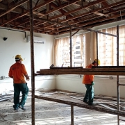 Uma fotografia colorida em plano geral e perspectiva ao n&iacute;vel dos olhos capta trabalhadores da constru&ccedil;&atilde;o civil durante as obras de reforma e recupera&ccedil;&atilde;o de uma sala na Escola Estadual Dem&eacute;trio Ribeiro, em Alegrete.

**Os Trabalhadores e a A&ccedil;&atilde;o (em primeiro e meio plano):**

* No centro e &agrave; esquerda da composi&ccedil;&atilde;o, dois oper&aacute;rios trabalham na desmontagem e remodela&ccedil;&atilde;o do espa&ccedil;o, vestindo camisas de trabalho de manga comprida de cor laranja-vibrante, cal&ccedil;as de prote&ccedil;&atilde;o verdes, capacetes de seguran&ccedil;a amarelos e botas de trabalho.


* O oper&aacute;rio da esquerda caminha de costas em dire&ccedil;&atilde;o &agrave; parede, segurando um rolo de fita adesiva/medida na m&atilde;o direita.


* O oper&aacute;rio da direita est&aacute; posicionado sobre a plataforma de t&aacute;buas de madeira de um andaime met&aacute;lico, trabalhando na remo&ccedil;&atilde;o de materiais do teto.


* Em primeiro plano, destaca-se a estrutura tubular met&aacute;lica em tom castanho/enferrujado de um andaime de dois n&iacute;veis.



**A Estrutura e o Ambiente da Sala:**

* No teto, exp&otilde;e-se a estrutura de ripas de madeira do telhado ap&oacute;s a remo&ccedil;&atilde;o do forro original, com telhas onduladas vis&iacute;veis no topo.


* As paredes est&atilde;o pintadas de branco/creme, com tubagens el&eacute;tricas externas de alum&iacute;nio e tomadas instaladas a meia altura. Na parede do fundo, sobressai um aparelho de ar condicionado do tipo *split* branco.


* Ao fundo, grandes janelas est&atilde;o cobertas por persianas verticais de tecido bege/castanho-claro, filtrando a luz solar intensa do exterior.


* O ch&atilde;o de cer&acirc;mica clara est&aacute; repleto de restos de madeira, ripas e entulho resultantes da desmontagem da cobertura.