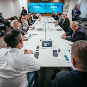 Uma fotografia colorida em plano geral e perspectiva em contra-campo (em &acirc;ngulo ligeiramente elevado) capta uma reuni&atilde;o de trabalho e alinhamento sobre a transi&ccedil;&atilde;o de governo na &aacute;rea da sa&uacute;de.

**As Autoridades e a Mesa de Reuni&atilde;o (ao centro e em primeiro plano):**

* Em primeiro plano, no centro inferior da imagem, observam-se as costas de dois participantes sentados &agrave; extremidade de uma grande mesa retangular de confer&ecirc;ncias de cor branca. O participante da esquerda veste camisa social branca e o da direita veste blazer escuro.


* Sobre o tampo da mesa, ao centro, destaca-se um tablet de controlo de videoconfer&ecirc;ncia, juntamente com telem&oacute;veis, comandos remotos, copos de &aacute;gua, blocos de notas, canetas e computadores port&aacute;teis abertos.


* Ao longo das duas extensas laterais da mesa, alinham-se cerca de uma dezena e meia de gestores, secret&aacute;rios e t&eacute;cnicos em trajes formais e esporte fino (blazers, camisas e casacos):


* &Agrave; esquerda, destaca-se a secret&aacute;ria estadual da Sa&uacute;de, Arita Bergmann, ao lado de gestoras que analisam relat&oacute;rios e documentos impressos.


* &Agrave; direita, alinham-se outros integrantes das equipas de transi&ccedil;&atilde;o consultando computadores e apontamentos.





**Os Monitores de Apresenta&ccedil;&atilde;o e o Ambiente (ao fundo):**

* Ao fundo da sala, centrados na parede principal, sobressaem dois grandes ecr&atilde;s/monitores de televis&atilde;o alinhados lado a lado transmitindo a apresenta&ccedil;&atilde;o oficial com o t&iacute;tulo: **"TRANSI&Ccedil;&Atilde;O DE GOVERNO"** sobre fundo azul-celeste.


* Ao fundo e nas extremidades da sala, outros assessores e profissionais de imprensa acompanham o encontro em p&eacute; ou dispostos em cadeiras de apoio.


* O ambiente conta com paredes em tom creme/bege claro, piso revestido a alcatifa cinzenta e ilumina&ccedil;&atilde;o embutida uniforme no teto.
