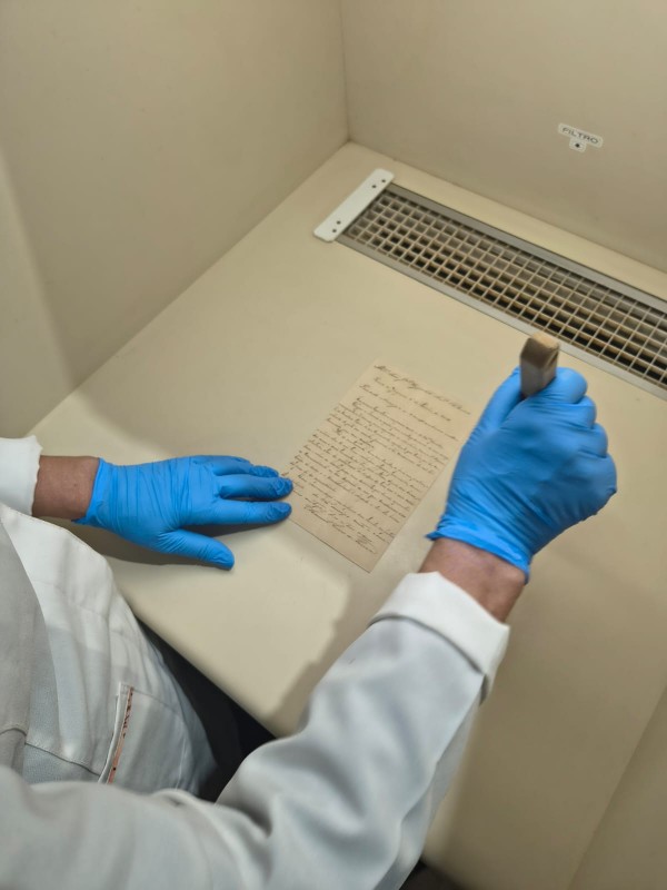 Foto mostra o detalhe das mãos de um técnico, com jaleco branco e luvas azuis, em frente uma bancada bege onde está sendo analisada uma carta antiga manuscrita. 