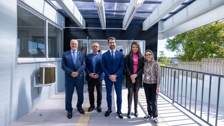 Grupo de pessoas posando em uma passarela coberta de um edif&iacute;cio moderno, com paredes em tons claros, cobertura transl&uacute;cida e guarda-corpo met&aacute;lico; ao fundo, h&aacute; portas envidra&ccedil;adas e vista parcial da &aacute;rea externa.
