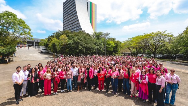 Uma fotografia colorida em plano geral e perspetiva em contra-plong&eacute;e suave (vista de &acirc;ngulo elevado) capta uma grande foto coletiva de servidores p&uacute;blicos estaduais reunidos em apoio &agrave; campanha do Outubro Rosa no p&aacute;tio do Centro Administrativo Fernando Ferrari (CAFF), em Porto Alegre.

**Os Servidores e o Movimento Outubro Rosa (em primeiro e meio plano):**

* Em primeiro plano e no centro da composi&ccedil;&atilde;o, uma multid&atilde;o formada por dezenas de servidores p&uacute;blicos pose alinhada e sorridente para a fotografia oficial.


* A grande maioria dos participantes veste pe&ccedil;as de vestu&aacute;rio em tons de cor-de-rosa (t-shirts, blusas, casacos, blazers e cal&ccedil;as) e muitos seguram pequenas rosas cor-de-rosa ou brancas nas m&atilde;os.


* No centro do grupo da frente, destaca-se a secret&aacute;ria estadual da Sa&uacute;de, Arita Bergmann, acompanhada por gestores e lideran&ccedil;as da pasta.


* O grupo encontra-se reunido sobre o pavimento intertravado de bet&atilde;o (paver) de uma grande esplanada exterior.



**O Edif&iacute;cio do CAFF e o Entorno (ao fundo):**

* Ao fundo, no centro superior da imagem, ergue-se o ic&oacute;nico edif&iacute;cio principal do Centro Administrativo Fernando Ferrari (CAFF), com a sua arquitetura vertical curva caracter&iacute;stica e um painel de grandes dimens&otilde;es nas cores da bandeira do Rio Grande do Sul (verde, vermelho e amarelo) verticais na sua fachada.


* A ladear o edif&iacute;cio e em redor da esplanada, observam-se &aacute;rvores frondosas com folhagem verde intensa.


* &Agrave; esquerda, avista-se uma rampa de acesso ao complexo e ve&iacute;culos estacionados.


* O c&eacute;u apresenta-se num tom azul-claro com nuvens brancas e esparsas, sob ilumina&ccedil;&atilde;o solar direta de dia claro.
