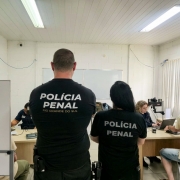Foto mostra dois policiais penais, de costa, com camisetas pretas da Pol&iacute;cia Penal, na frente de uma sala onde atendentes est&atilde;o coletando os dados dos detentos. 