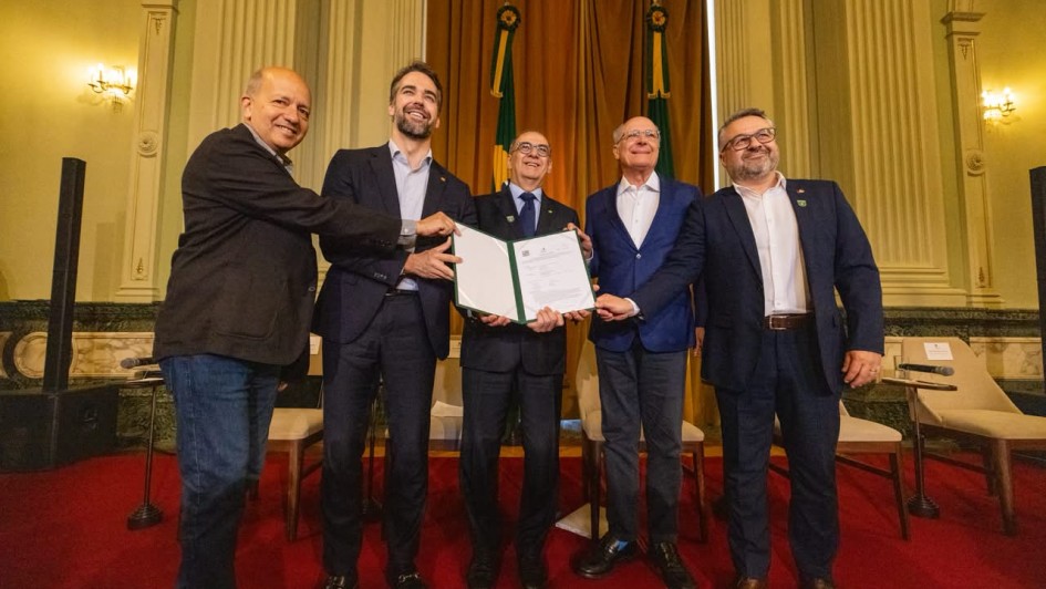 Grupo de pessoas posa sobre o palco exibindo um documento aberto para a c&acirc;mera, durante uma cerim&ocirc;nia oficial realizada em um sal&atilde;o hist&oacute;rico com bandeiras ao fundo e tapete vermelho.