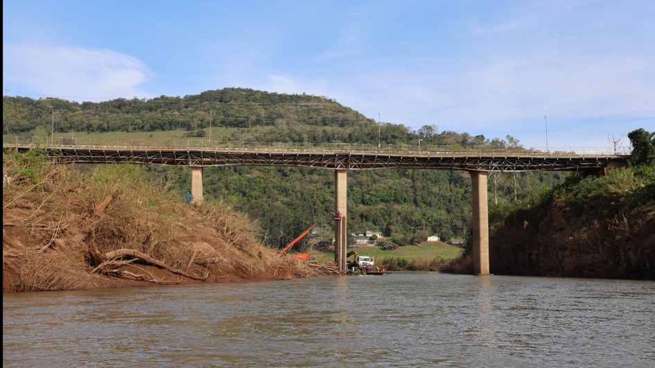 Foto mostra o rio com a ponte ao fundo. Atr&aacute;s, aparece um morro verde. Na margem esquerda, h&aacute; ra&iacute;zes expostas, provavelmente arrastadas pela enchente. 