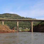 Foto mostra o rio com a ponte ao fundo. Atr&aacute;s, aparece um morro verde. Na margem esquerda, h&aacute; ra&iacute;zes expostas, provavelmente arrastadas pela enchente. 