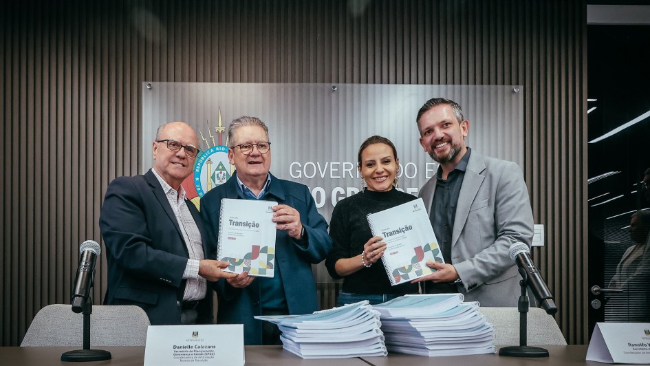 Uma fotografia colorida em plano m&eacute;dio-geral e perspectiva ao n&iacute;vel dos olhos capta quatro autoridades estaduais apresentando os Livros de Transi&ccedil;&atilde;o oficializados para subsidiar a pr&oacute;xima gest&atilde;o governamental.

**As Autoridades e os Livros de Transi&ccedil;&atilde;o (ao centro e em primeiro plano):**

* No centro da composi&ccedil;&atilde;o, quatro gestores posam alinhados lado a lado atr&aacute;s de uma mesa de confer&ecirc;ncias:
* &Agrave; esquerda, um homem de cabelos grisalhos curtos e &oacute;culos veste um blazer azul-escuro sobre camisa clara e junta-se ao colega ao lado para segurar um exemplar encadernado.
* Ao seu lado, outro gestor de cabelos grisalhos e &oacute;culos segura com ambas as m&atilde;os o livro de capa clara com espiral branca onde se l&ecirc; em destaque a palavra **"Transi&ccedil;&atilde;o"** acompanhada de grafismos geom&eacute;tricos coloridos.
* &Agrave; sua direita, a secret&aacute;ria de Planejamento, Governan&ccedil;a e Gest&atilde;o, Danielle Calazans, sorri vestindo blusa escura de mangas compridas e segura outro exemplar do Livro de Transi&ccedil;&atilde;o com o mesmo grafismo institucional.
* Na extremidade direita, um gestor de barba e cabelo curto veste blazer cinzento-claro sobre camisa escura, segurando o exemplar ao lado da secret&aacute;ria.


* Em primeiro plano, sobre o tampo da mesa, empilham-se dezenas de relat&oacute;rios e documentos oficiais encadernados em espiral branca.
* Nas extremidades da mesa, destacam-se dois microfones com base de mesa e identificadores em acr&iacute;lico com placas nominais.

**O Painel do Fundo e o Ambiente (ao fundo):**

* Ao fundo, a parede de fundo &eacute; revestida por ripas verticais de madeira em tom castanho-m&eacute;dio.
* No centro da parede, sobressai um painel acr&iacute;lico transparente com o bras&atilde;o oficial do Estado do Rio Grande do Sul e as letras mai&uacute;sculas em relevo met&aacute;lico de parte do texto institucional: **"GOVERNO DO ESTADO"**.
* A ilumina&ccedil;&atilde;o no interior do espa&ccedil;o &eacute; limpa e focada sobre as autoridades, destacando o ato solene de entrega dos documentos.