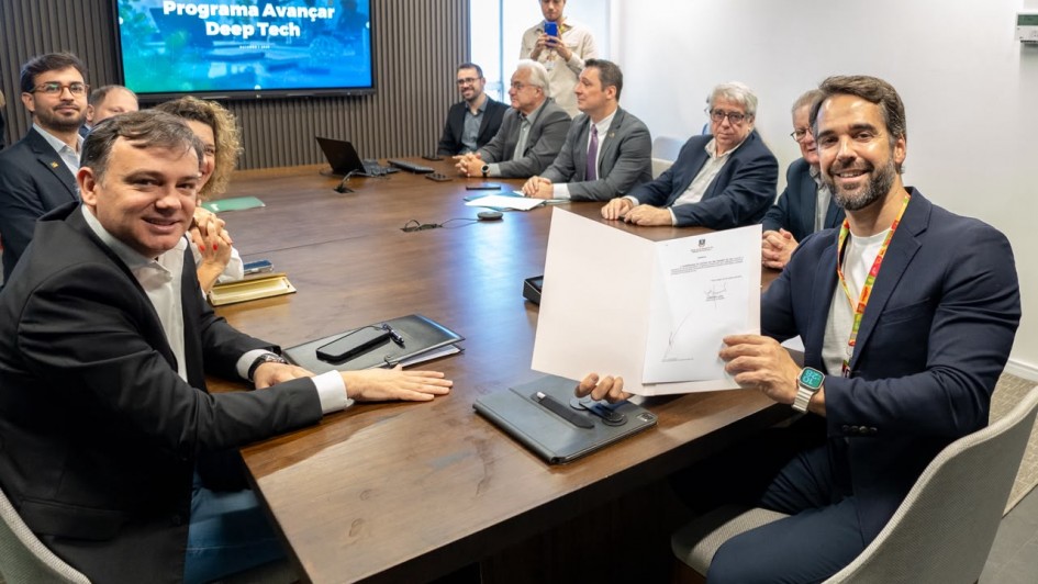 Uma fotografia colorida em plano m&eacute;dio e perspectiva em &acirc;ngulo levemente elevado capta o governador Eduardo Leite e gestores estaduais durante o ato de assinatura do decreto de institui&ccedil;&atilde;o do Programa Avan&ccedil;ar Deep Tech.

**As Autoridades e a Assinatura (em primeiro plano):**

* Em primeiro plano, &agrave; direita, o governador Eduardo Leite sorri para a c&acirc;mara enquanto segura com ambas as m&atilde;os uma pasta com o decreto oficial assinado. Ele veste um blazer azul-marinho sobre camisola de gola redonda clara, fita/cord&atilde;o institucional ao pesco&ccedil;o e rel&oacute;gio digital com mostrador verde no pulso esquerdo.
* &Agrave; esquerda, em primeiro plano, um gestor sorri virado para a c&acirc;mara, trajando blazer escuro e camisa social branca de colarinho aberto, com o bra&ccedil;o esquerdo apoiado sobre uma pasta de couro na mesa.
* Ambos est&atilde;o sentados em cadeiras de escrit&oacute;rio estofadas em tom cinzento-claro nas extremidades de uma grande mesa de reuni&otilde;es retangular de madeira escura.

**A Mesa de Reuni&atilde;o e o Painel (ao fundo):**

* Ao longo do lado oposto da mesa, alinham-se cerca de meia dezena de secret&aacute;rios, diretores e t&eacute;cnicos em trajes formais (fatos, blazers e gravatas), acompanhando atentamente o ato de assinatura.
* Ao fundo, &agrave; esquerda, sobressai um ecr&atilde; de televis&atilde;o fixado numa parede com pain&eacute;is de madeira ripada vertical. O ecr&atilde; exibe a apresenta&ccedil;&atilde;o oficial com o cabe&ccedil;alho **"ASSINATURA DE DECRETO"** e o t&iacute;tulo principal em letras brancas: **"Programa Avan&ccedil;ar Deep Tech"** sobre fundo azul tecnol&oacute;gico.
* Ao fundo e ao centro, um assessor em p&eacute; regista o momento com um smartphone.
* O ambiente conta com ilumina&ccedil;&atilde;o interior clara e uniforme, transmitindo um clima solene e institucional.