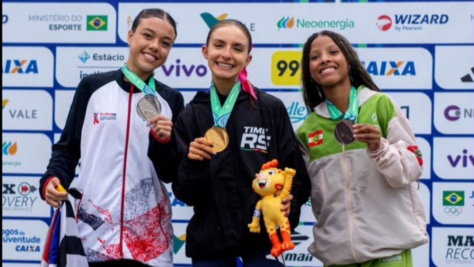 Foto mostra atleta Emanueli Maiara Hein, ao centro, com outras duas atletas. Elas mostram suas medalhas. Ela est&aacute; com um abrigo esportivo preto com o dizer Times RS.