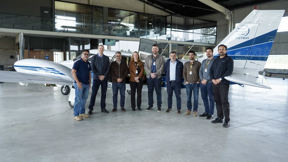 Uma fotografia colorida em plano geral e perspectiva ao n&iacute;vel dos olhos regista uma comitiva de t&eacute;cnicos, gestores e especialistas reunidos num hangar aeroportu&aacute;rio para o avan&ccedil;o do mapeamento topogr&aacute;fico do territ&oacute;rio do Rio Grande do Sul.

**O Grupo e as Autoridades (ao centro e em primeiro plano):**

* No centro da composi&ccedil;&atilde;o, nove pessoas posam alinhadas lado a lado em p&eacute; sobre o piso de cimento polido e cinzento do hangar.
* O grupo veste trajes esporte fino e casuais (casacos, blazers, camisas polo, camisas sociais e cal&ccedil;as jeans/escuras) e v&aacute;rios membros utilizam fita/cord&atilde;o institucional de crach&aacute; ao pesco&ccedil;o.


* &Agrave; esquerda, destaca-se um integrante com t-shirt polo azul-marinho e cal&ccedil;as jeans. Ao centro, observam-se gestores de casaco castanho, jaqueta e blazer azul-escuro.


* &Agrave; direita, alinham-se t&eacute;cnicos e especialistas com camisas sociais, camisolas e jaquetas escuras, com as m&atilde;os cruzadas &agrave; frente do corpo.



**A Aeronave e a Infraestrutura do Hangar (ao fundo):**

* Logo atr&aacute;s do grupo, sobressai uma aeronave bimotor de aerofotogrametria de cor predominantemente branca com detalhes e faixas em tons de azul-claro e azul-escuro na fuselagem e na cauda/estabilizador vertical.


* Na cauda do avi&atilde;o, l&ecirc;-se a marca **"ESTEIO"** acompanhada de um log&oacute;tipo circular.


* Ao fundo, destaca-se a estrutura moderna do hangar, com mezanino envidra&ccedil;ado de dois pisos, caixilharia met&aacute;lica escura, teto com vigas de a&ccedil;o e ampla ilumina&ccedil;&atilde;o natural difusa vinda das portas abertas do pavilh&atilde;o.