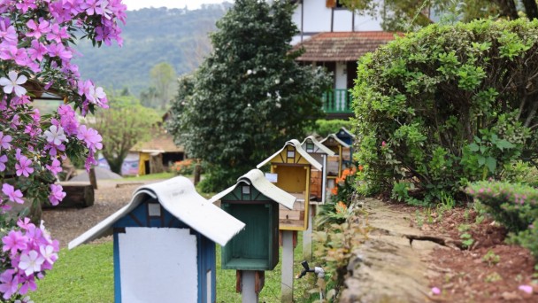 Imagem mostra parte de uma casa em estilo colonial ao fundo e no primeiro plano seis casinhas estilizadas de cores diferentes, como se fossem caixas de abelhas