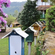 Imagem mostra parte de uma casa em estilo colonial ao fundo e no primeiro plano seis casinhas estilizadas de cores diferentes, como se fossem caixas de abelhas
