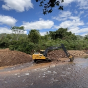 imagem mostra uma m&aacute;quina escavadeira retirando sedimentos de um rio raso.