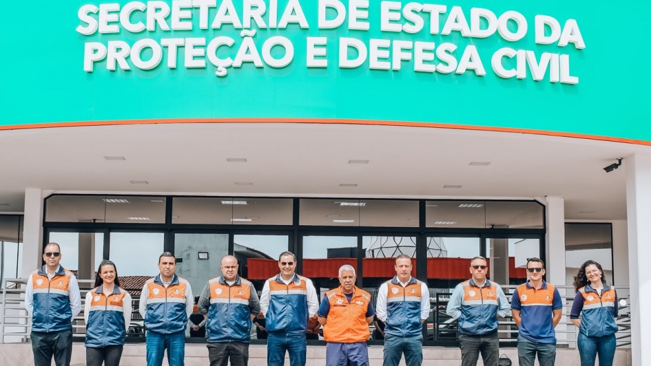 Des pessoas estao lado a lado na frente da entrada de um pr&eacute;dio onde tem a inscri&ccedil;&atilde;o Secretaria de Estado da Prote&ccedil;&atilde;o e Defesa Civil