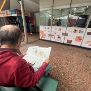 Imagem mostra um homem em primeiro plano lendo um folheto sentado em frente a um estante da Secretaria da Cultura do Rio Grande do Sul na Feira do Livro de Porto Alegre 